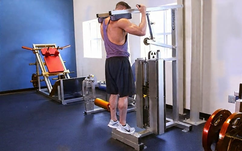 Man performing standing calf raise exercise on a standing calf raise machine in a gym, wearing a blue tank top and black shorts, with a window and other gym equipment in the background.